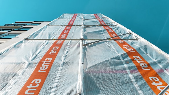 A tall building covered in scaffolding with protective sheeting. The sheeting has a repeating orange stripe with the word 'renta' on it. The building is set against a clear blue sky.