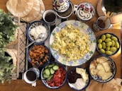 An assortment of foods arranged on a wooden table, featuring scrambled eggs with herbs on a decorative plate surrounded by side dishes including yogurt, olives, tomato and cucumber slices, a spiced eggplant dish, pita bread, and different spreads. Tea is being poured from a pot into a cup. Potted plants add a touch of greenery to the scene.