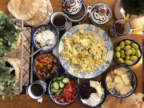 An assortment of foods arranged on a wooden table, featuring scrambled eggs with herbs on a decorative plate surrounded by side dishes including yogurt, olives, tomato and cucumber slices, a spiced eggplant dish, pita bread, and different spreads. Tea is being poured from a pot into a cup. Potted plants add a touch of greenery to the scene.