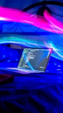 A laptop displaying code on its screen is placed on a bed with a blue bedsheet. In the background, vibrant pink and blue light trails create a dynamic and futuristic atmosphere.