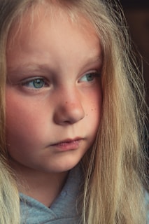 A close-up portrait of a young girl with long blonde hair and blue eyes. Her face is shown in profile, and she appears deep in thought. The lighting is soft and natural, highlighting her facial features.