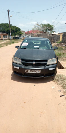 A black car is parked on a dirt road. The car has a sign in the windshield and the side window. The background features a residential area with houses, trees, and a brick wall. The road is unpaved, and there are utility poles and wires above.