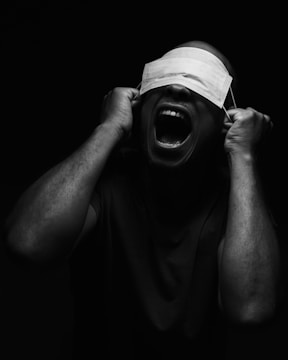 A person is holding a face mask over their eyes with their mouth wide open, set against a dark background.