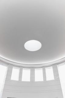 A minimalist architectural interior featuring a circular dome with a central round skylight. The surrounding area consists of evenly spaced vertical windows, allowing natural light to fill the space. The walls and ceiling are smooth and predominantly white, contributing to a clean and modern aesthetic.