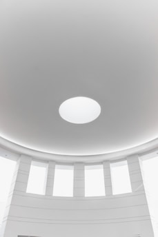 A minimalist architectural interior featuring a circular dome with a central round skylight. The surrounding area consists of evenly spaced vertical windows, allowing natural light to fill the space. The walls and ceiling are smooth and predominantly white, contributing to a clean and modern aesthetic.