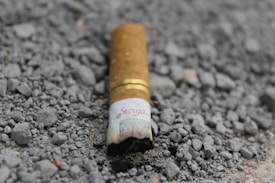 A cigarette butt partially burnt lies discarded on a rough, gravel-covered ground. The brand name is visible on the filter paper.