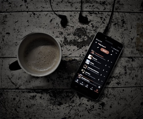 A rustic wooden surface displays a cup of coffee next to a Samsung smartphone with earphones. The phone screen shows a music playlist in a dark-themed music app, with the song 'Friendship Avenue' highlighted. The dim and moody lighting adds to the ambience.
