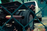 A white truck with missing parts, notably the bed and rear coverings, is parked in a lot. The image is taken through a chain-link fence, giving a sense of separation or confinement. The truck appears to be in a state of disrepair, with visible rust on the metal frame and exhaust. The background features a blue building with an air conditioning unit in the window, and a small plant is visible in the lower part of the image.