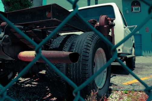 A white truck with missing parts, notably the bed and rear coverings, is parked in a lot. The image is taken through a chain-link fence, giving a sense of separation or confinement. The truck appears to be in a state of disrepair, with visible rust on the metal frame and exhaust. The background features a blue building with an air conditioning unit in the window, and a small plant is visible in the lower part of the image.