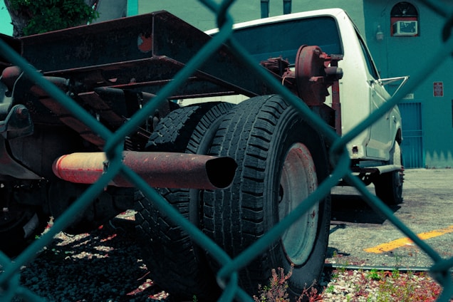A white truck with missing parts, notably the bed and rear coverings, is parked in a lot. The image is taken through a chain-link fence, giving a sense of separation or confinement. The truck appears to be in a state of disrepair, with visible rust on the metal frame and exhaust. The background features a blue building with an air conditioning unit in the window, and a small plant is visible in the lower part of the image.