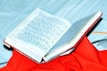 An open book with text in a script that resembles Hindi or Sanskrit, placed on top of a vibrant red cloth. The book is partially open, displaying printed content, and is set against a light blue background.