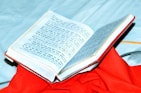 An open book with text in a script that resembles Hindi or Sanskrit, placed on top of a vibrant red cloth. The book is partially open, displaying printed content, and is set against a light blue background.