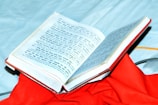 An open book with text in a script that resembles Hindi or Sanskrit, placed on top of a vibrant red cloth. The book is partially open, displaying printed content, and is set against a light blue background.