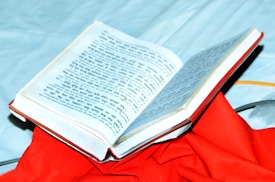 An open book with text in a script that resembles Hindi or Sanskrit, placed on top of a vibrant red cloth. The book is partially open, displaying printed content, and is set against a light blue background.
