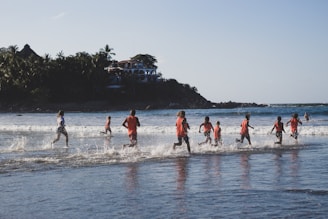 A group of children wearing red shirts are joyfully running through shallow ocean waves near a serene beach. In the background, lush greenery and a hillside dotted with buildings provide a picturesque setting. The sun casts a warm glow, enhancing the cheerful atmosphere.