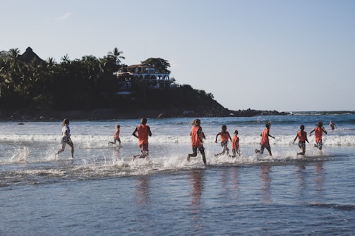 A group of children wearing red shirts are joyfully running through shallow ocean waves near a serene beach. In the background, lush greenery and a hillside dotted with buildings provide a picturesque setting. The sun casts a warm glow, enhancing the cheerful atmosphere.