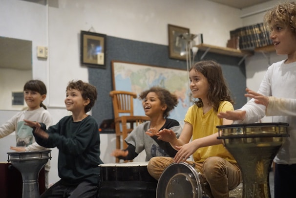 Five children are joyfully playing drums in a warmly lit room. The children, each seated in a row, appear to be fully engaged and smiling as they beat on various percussion instruments. The background has a map, a wooden chair, and a few wall decorations.