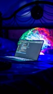 A laptop displaying code is placed on a bed with a vibrant, colorful projection or light installation in the background. The scene is dimly lit with a strong emphasis on blues and neon colors, creating a futuristic and technological atmosphere.