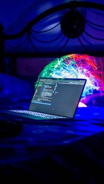 A laptop displaying code is placed on a bed with a vibrant, colorful projection or light installation in the background. The scene is dimly lit with a strong emphasis on blues and neon colors, creating a futuristic and technological atmosphere.