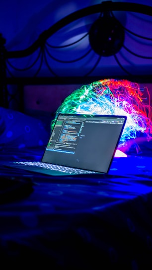 A laptop displaying code is placed on a bed with a vibrant, colorful projection or light installation in the background. The scene is dimly lit with a strong emphasis on blues and neon colors, creating a futuristic and technological atmosphere.