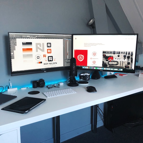 A modern workspace featuring a dual-monitor setup on a white desk. The screens display graphic design work and a webpage. Various accessories like a keyboard, mouse, microphone, camera, and a tablet are neatly arranged. The ambiance is enhanced with subtle blue lighting.