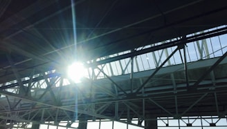 Sunlight shines brightly through the steel beams and skylights of a large industrial or commercial structure. The beams create a pattern of intersecting lines while the sunlight casts a glowing aura and lens flare effect.