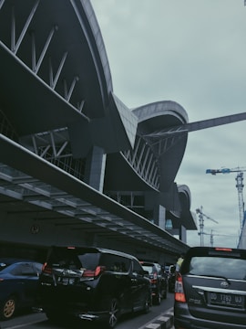 The image captures a modern architectural structure with curved lines and steel details. Several vehicles are visible in the foreground, possibly indicating a busy area or a drop-off zone. Construction cranes can be seen in the background, suggesting ongoing developments in the vicinity.