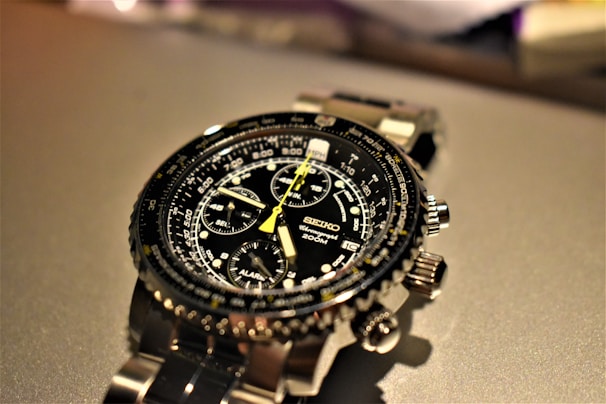 A detailed wristwatch with a black dial featuring multiple subdials and a yellow second hand. The watch has a stainless steel strap and various markings and labels, including '200M' and 'Seiko'. The background is out of focus, drawing attention to the watch.