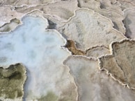 Aerial view of natural salt formations with intricate patterns, featuring a mix of textures and colors including pale blue and beige. The formations are irregular in shape and spread across the surface, resembling terraced structures with mineral deposits along the edges.