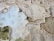 Aerial view of natural salt formations with intricate patterns, featuring a mix of textures and colors including pale blue and beige. The formations are irregular in shape and spread across the surface, resembling terraced structures with mineral deposits along the edges.