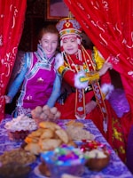 Two people dressed in vibrant traditional attire stand beside a table filled with a variety of foods. The individuals wear elaborate costumes with intricate designs and bold colors. One person wears a headdress featuring red, blue, and gold ornamentation. The background is adorned with red curtains, adding to the festive atmosphere.