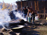 Two people are cooking outdoors in large pots over an open fire, with smoke rising and a rustic wooden building in the background. There are trees in the vicinity, indicating an outdoor setting, possibly in a forest or rural area.