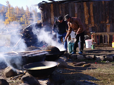 Two people are cooking outdoors in large pots over an open fire, with smoke rising and a rustic wooden building in the background. There are trees in the vicinity, indicating an outdoor setting, possibly in a forest or rural area.
