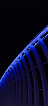 A series of curved vertical beams are illuminated with a rich blue light against a dark background. The architectural design creates a repetitive and modern pattern that suggests a building or a structure. The blue lighting contrasts sharply with the dark sky, emphasizing the curvature and precision of the beams.