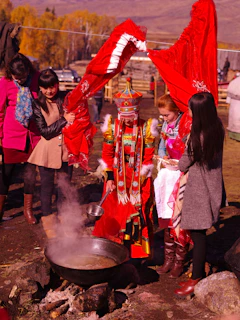 A group of people are gathered around a pot outdoors. They are dressed in traditional, vibrant clothing, with one individual wearing a particularly elaborate outfit made of red and multicolored patterns, holding a ladle. The scene appears to be cultural or ceremonial, with one person assisting in lifting a large red cloth.