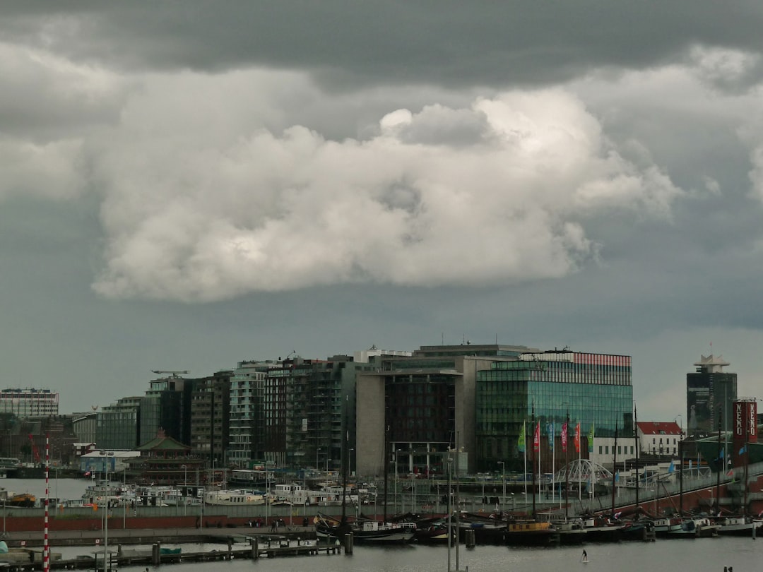 white and blue boat on body of water near city buildings under white clouds during daytime, New architecture picture in Amsterdam - wide city-view on the modern buildings of the Oosterdokseiland area in town center, under a cloudy and grey summer sky above the city. This photo shows the characteristic Dutch grey weather and its Dutch light. This image shows recently-built architecture of residential blocks and shops on the ground floor - all located along the wide Oosterdok water.<p></figure>Amsterdam city in free photos - urban photography of The Netherlands by Fons Heijnsbroek, pictures of Dutch modern architecture in town, 21 July 2012 