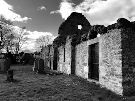 A black and white photograph of an ancient stone structure, possibly a ruin, with arched openings and remnants of walls. The setting appears to be a graveyard with several tombstones in the grassy foreground. Bare trees are seen in the background under a partly cloudy sky, creating a somber and historical atmosphere.
