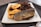 A plate holds a serving of fried plantains, a piece of grilled or roasted fish, and a pile of what appears to be rice mixed with spices and possibly vegetables. The dish is presented on a square ceramic plate set on a dark tray, with a spoon visible in the foreground.