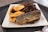 A plate holds a serving of fried plantains, a piece of grilled or roasted fish, and a pile of what appears to be rice mixed with spices and possibly vegetables. The dish is presented on a square ceramic plate set on a dark tray, with a spoon visible in the foreground.