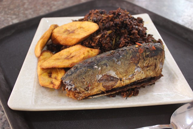 A plate holds a serving of fried plantains, a piece of grilled or roasted fish, and a pile of what appears to be rice mixed with spices and possibly vegetables. The dish is presented on a square ceramic plate set on a dark tray, with a spoon visible in the foreground.