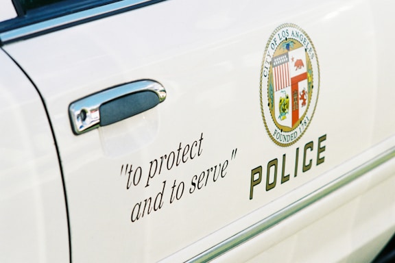 The side of a white police car features the motto 'to protect and to serve' in black script near the door handle. The emblem of the City of Los Angeles with various symbols is also displayed next to the word 'POLICE' in bold letters.