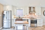 A modern kitchen with white cabinetry and a metallic refrigerator located to the left. The central area features a farmhouse sink under a window, flanked by countertops. A stainless steel range with a backsplash made of light brown bricks is topped by an exhaust hood. A kitchen island with a wooden top is placed at the center, while a wall clock is visible on the right.
