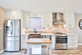 A modern kitchen with white cabinetry and a metallic refrigerator located to the left. The central area features a farmhouse sink under a window, flanked by countertops. A stainless steel range with a backsplash made of light brown bricks is topped by an exhaust hood. A kitchen island with a wooden top is placed at the center, while a wall clock is visible on the right.