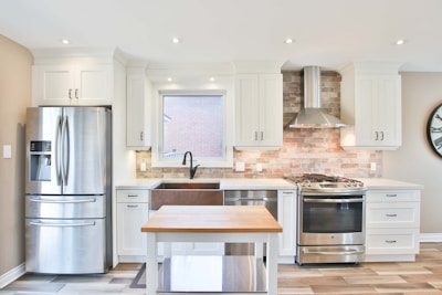 A modern kitchen with white cabinetry and a metallic refrigerator located to the left. The central area features a farmhouse sink under a window, flanked by countertops. A stainless steel range with a backsplash made of light brown bricks is topped by an exhaust hood. A kitchen island with a wooden top is placed at the center, while a wall clock is visible on the right.