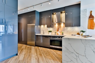A modern kitchen with sleek gray cabinets, stainless steel appliances, and a marble countertop. There is a wooden cutting board hanging on the wall, recessed lighting, and decorative plants placed on the counter. The wooden flooring adds warmth to the contemporary space.