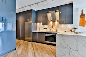 A modern kitchen with sleek gray cabinets, stainless steel appliances, and a marble countertop. There is a wooden cutting board hanging on the wall, recessed lighting, and decorative plants placed on the counter. The wooden flooring adds warmth to the contemporary space.