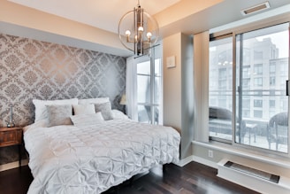 A cozy and stylish bedroom featuring a large, plush bed with white textured bedding and multiple pillows against a decorative gray damask-patterned wallpaper. A wooden side table with a minimalist lamp and a hanging modern chandelier provide lighting. The room is brightened by a large window with a view of adjacent buildings, adding a touch of urban elegance.