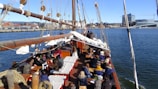 A large group of people is seated on the deck of a wooden sailing ship, which is in a harbor. Various white and beige ropes and sails are visible, and the cityscape with buildings and cranes is seen in the background across the water under a clear blue sky.