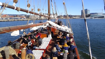 A large group of people is seated on the deck of a wooden sailing ship, which is in a harbor. Various white and beige ropes and sails are visible, and the cityscape with buildings and cranes is seen in the background across the water under a clear blue sky.
