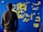 A person stands facing a blue wall covered with numerous yellow sticky notes arranged in a flowchart-like pattern. The notes are organized into categories with handwritten titles like 'Environment', 'Society', and 'Healthcare'.